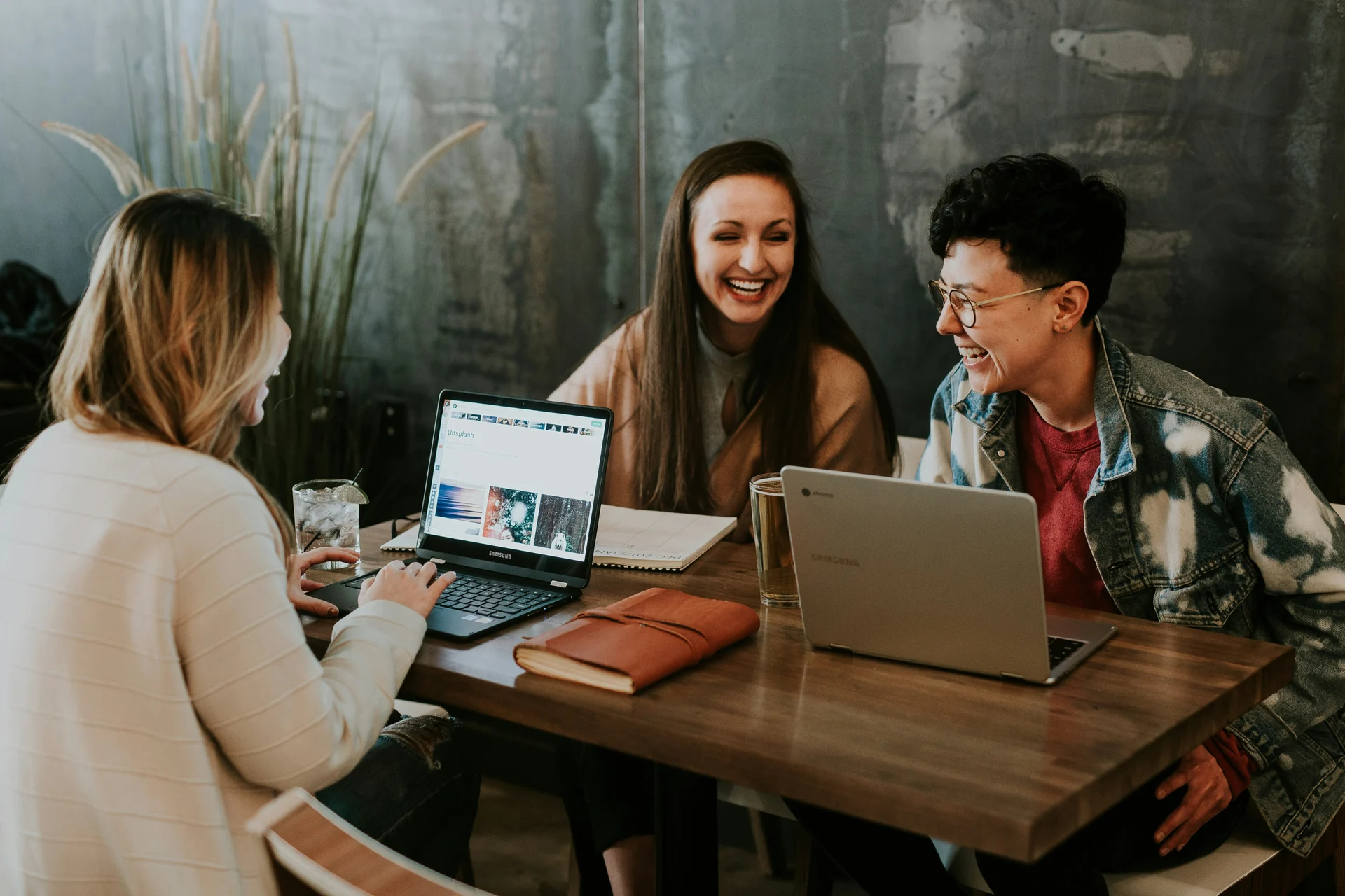 Three colleagues laughing over laptops at a shared table in a coworking space